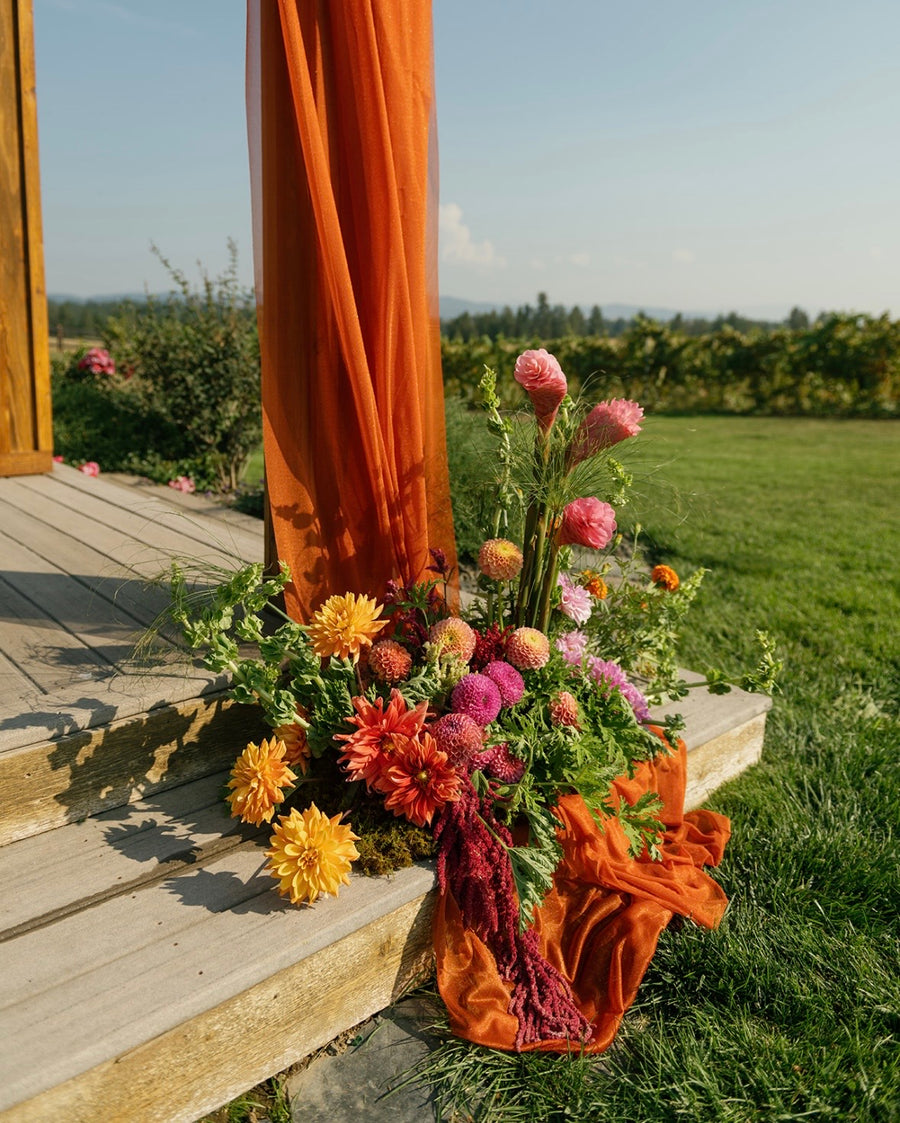 Floral arrangement with orange and pink flowers on a wooden deck with orange curtains.