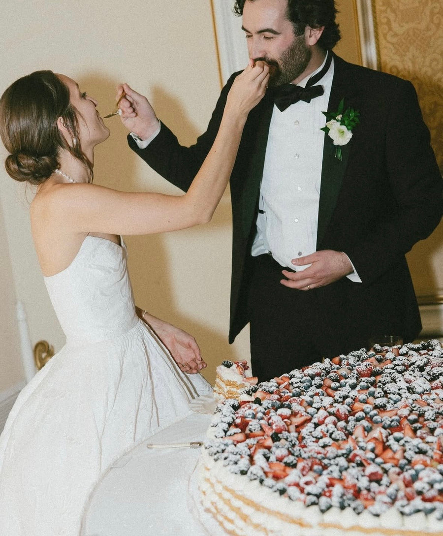 Man and woman in formal attire feeding each other at a wedding, with a decorated cake in the foreground.