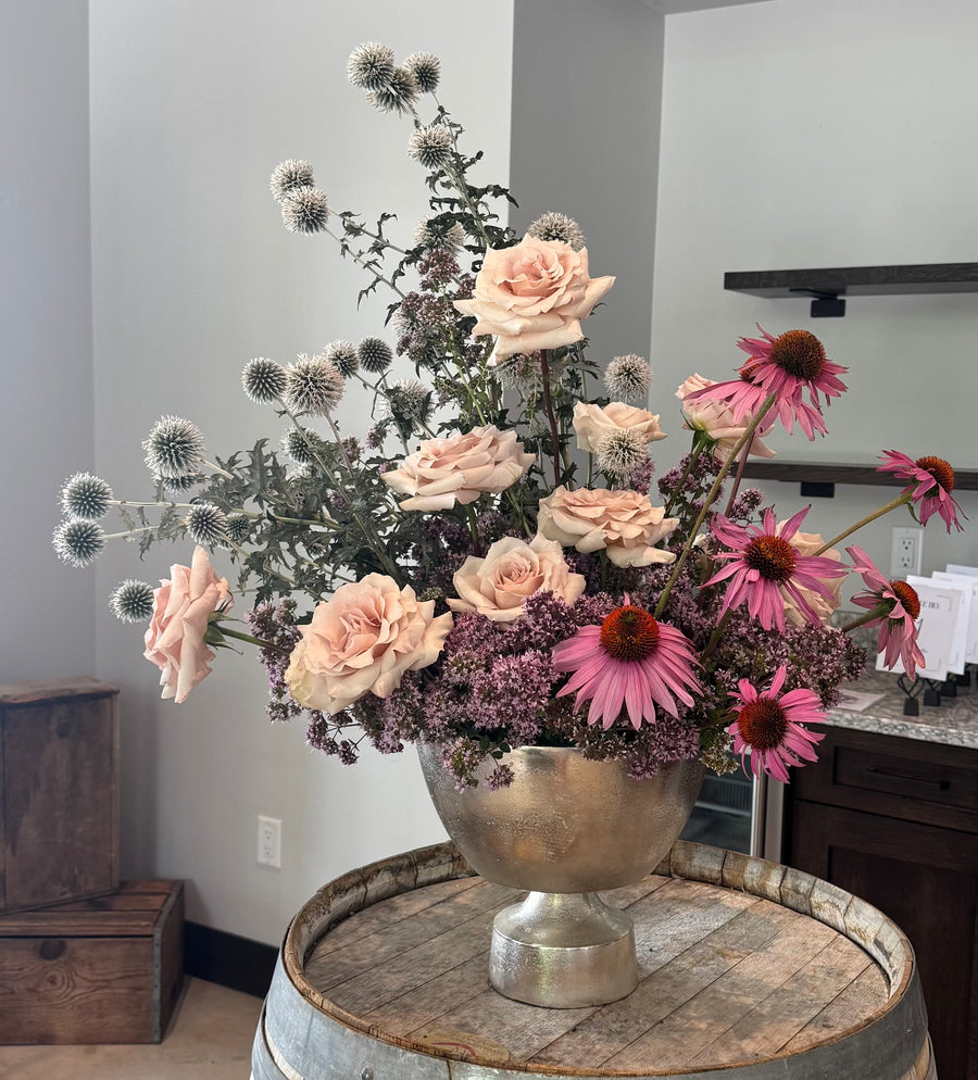 Floral arrangement in a silver vase on a wooden barrel in a room setting.